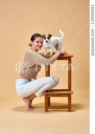 Portrait of female model posing with her lovely pet, purebred Jack Russell Terrier against beige background. Concept of pet lovers. 110099261