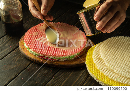 The cook is preparing a round waffle cake on the kitchen table. Chef adding condensed milk with spoon in hand The cook is preparing a round waffle cake on the kitchen table. Chef adding condensed milk with spoon in hand 110099500