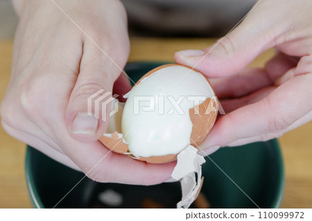A woman's hand peels a boiled egg, removing the shell close-up. A quick snack in the form of a boiled egg. Top view. 110099972