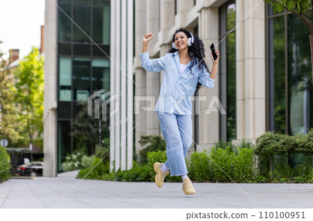 Young joyful beautiful woman dancing and singing in headphones while walking in the city, Hispanic woman with curly hair uses an application on the phone, to listen to music online. Young joyful beautiful woman dancing and singing in headphones while walking in the city, Hispanic woman with curly hair uses an application on the phone, to listen to music online. 110100951