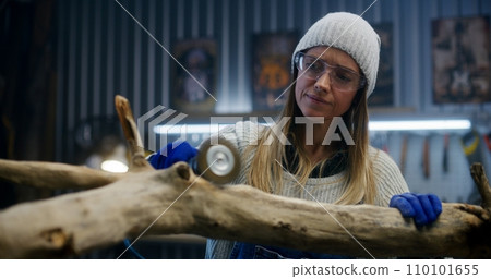 Female carpenter in goggles grinds tree branch using grinding machine 110101655