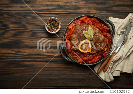 Traditional Italian dish Ossobuco all Milanese made with cut veal shank meat with vegetable tomato sauce served in black casserole pan top view on rustic brown wooden background, copy space Traditional Italian dish Ossobuco all Milanese made with cut veal shank meat with vegetable tomato sauce served in black casserole pan top view on rustic brown wooden background, copy space 110102982