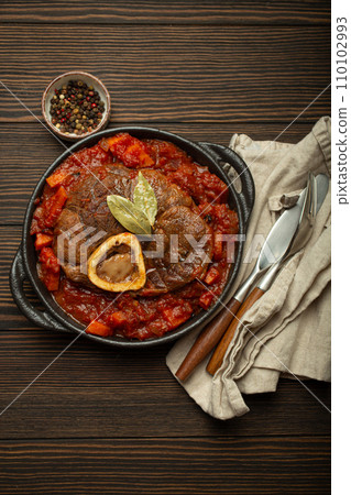 Traditional Italian dish Ossobuco all Milanese made with cut veal shank meat with vegetable tomato sauce served in black casserole pan top view on rustic brown wooden background 110102993