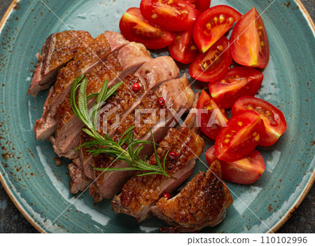 Delicious roasted sliced duck breast fillet with golden crispy skin, with pepper and rosemary, top view on ceramic blue plate served with cherry tomatoes salad, rustic concrete rustic background 110102996