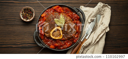 Traditional Italian dish Ossobuco all Milanese made with cut veal shank meat with vegetable tomato sauce served in black casserole pan top view on rustic brown wooden background 110102997
