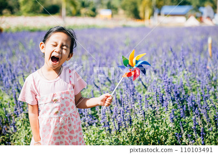 A happy girl stands in a flower garden with a toy pinwheel. Springtime's joy and the flying pinwheel evoke childhood fun happiness and freedom amidst the sunny nature-filled setting. 110103491