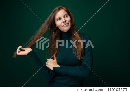 A young woman with brown hair stands thoughtfully on a dark green background 110105575