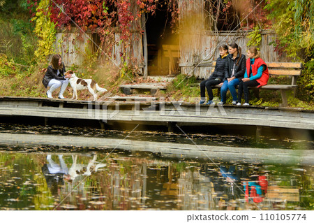 A young beautiful girl strokes a dog by the lake in an autumn park, a girl and two teenage girls are sitting next to her on a bench. 110105774