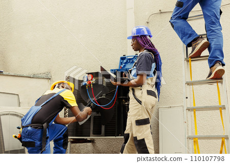 Seasoned mechanic cleaning and lubricating hvac system internal parts, checking refrigerant level and calibrating thermostats while african american colleague checks maintenance plan on laptop 110107798