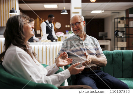 Caucasian elderly couple discussing their travel plans in luxury resort lounge area. Retired senior husband and wife waiting to check in at the hotel reception while planning a wonderful honeymoon. Caucasian elderly couple discussing their travel plans in luxury resort lounge area. Retired senior husband and wife waiting to check in at the hotel reception while planning a wonderful honeymoon. 110107853