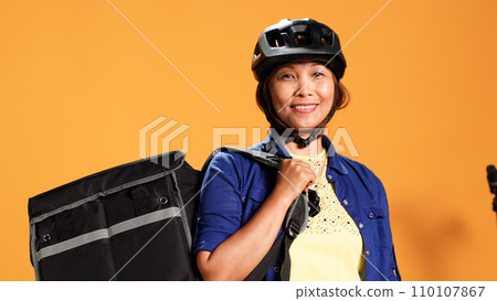 Portrait of happy asian courier using bike and thermic bag to transport perishables to customer. Delivery part time employee isolated over orange studio background wearing safety gear, close up 110107867