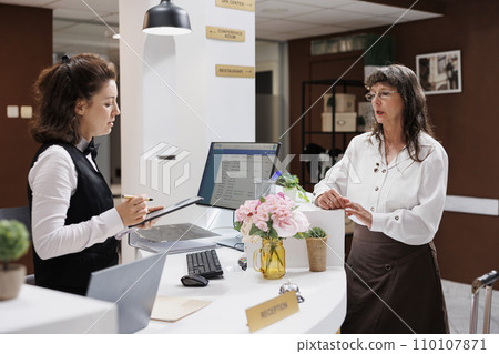 Experienced concierge helps elderly woman check in smoothly with digital device at hotel foyer. Front desk employee welcomes senior lady traveller and prepares reservation document on tablet. Experienced concierge helps elderly woman check in smoothly with digital device at hotel foyer. Front desk employee welcomes senior lady traveller and prepares reservation document on tablet. 110107871