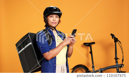 Portrait of courier waiting, looking in phone app for next customer order location. BIPOC woman delivering food with bycicle using thermic bag, isolated over studio background 110107875
