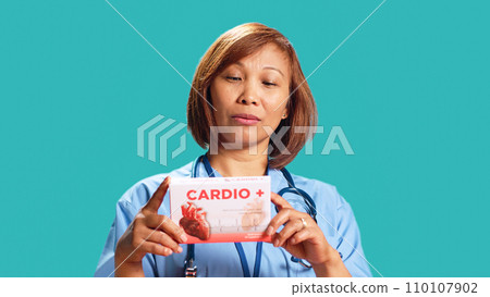 Healthcare professional holding miocardic tablets box, reading ingredients on pharmaceutical package, close up. Nurse isolated over studio background prescribing heart antibiotic to patient 110107902