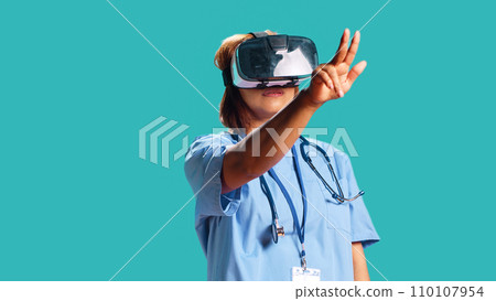 Healthcare professional wearing virtual reality headset remotely checking patient. Nurse isolated over blue studio background using high-tech advanced clinical vr gadget, close up Healthcare professional wearing virtual reality headset remotely checking patient. Nurse isolated over blue studio background using high-tech advanced clinical vr gadget, close up 110107954