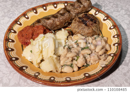 Closeup Plate With Traditional Bulgarian Food. Mashed Potato, White Beans, Haricot, Vegetable Relish Ljutenica, Grilled Minced Meat With Spices Kebapche. Horizontal Plane. Closeup Plate With Traditional Bulgarian Food. Mashed Potato, White Beans, Haricot, Vegetable Relish Ljutenica, Grilled Minced Meat With Spices Kebapche. Horizontal Plane. 110108485