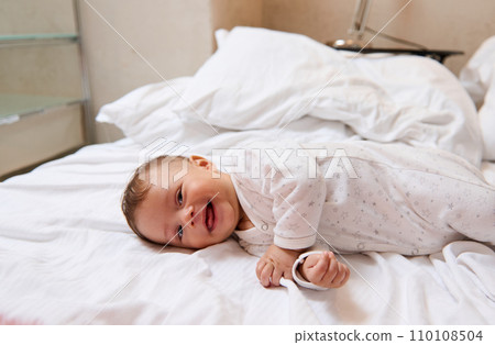 Playful lovely baby boy in white clothes, smiling and laughing lying on white bedsheets on the bed at home Playful lovely baby boy in white clothes, smiling and laughing lying on white bedsheets on the bed at home 110108504
