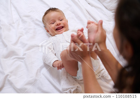 Beautiful newborn baby smiles looking at his loving mother, while she strokes and kisses his little feet and tiny toes 110108515