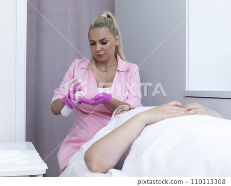 Esthetician Holds White Bottle with Peeling or Cleansing Foam, Cream above Beautiful Young Caucasian Female Patient. Beauty Procedure, Moisturizing Face Skin, Facial Massage. Horizontal 110113308