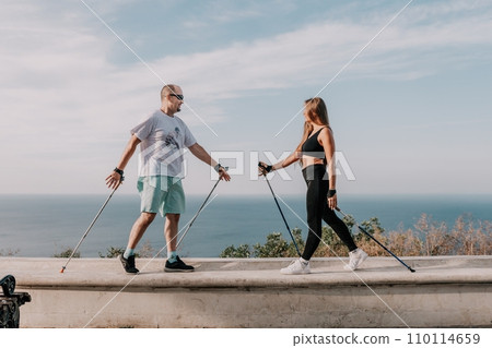 Happy Middle aged couple or friends practicing nordic walking in park near sea. Mature couple with trekking poles walking, practicing Nordic walking outdoors. Aging youthfully and sport concept 110114659
