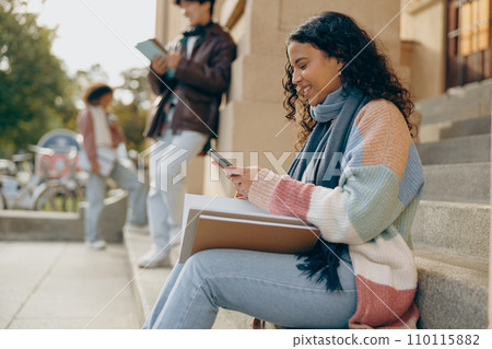 Smiling female student using phone sitting on stairs near university on friends background 110115882