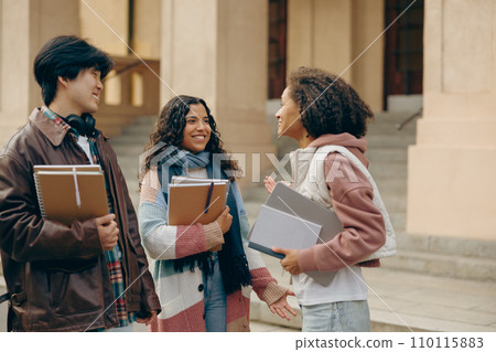 Young students talking and laughing together outdoor standing near campus and holding books Young students talking and laughing together outdoor standing near campus and holding books 110115883