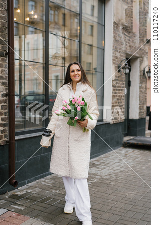 Young stylish woman with a takeaway coffee and a bouquet of tulips walking on a city street in springtime 110117240