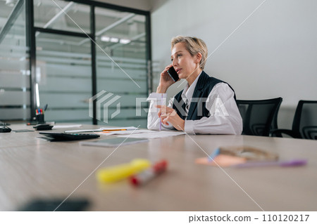 Low-angle view of elegant middle-aged businesswoman talking mobile phone with business partner in office. Mature thoughtful woman calling phone. Female entrepreneur having conversation on cellphone Low-angle view of elegant middle-aged businesswoman talking mobile phone with business partner in office. Mature thoughtful woman calling phone. Female entrepreneur having conversation on cellphone 110120217