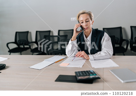 Portrait of middle-aged businesswoman making business call sitting on office desk, working with document. Front view of gray-haired female manager talking at smartphone with client discussing contract Portrait of middle-aged businesswoman making business call sitting on office desk, working with document. Front view of gray-haired female manager talking at smartphone with client discussing contract 110120218