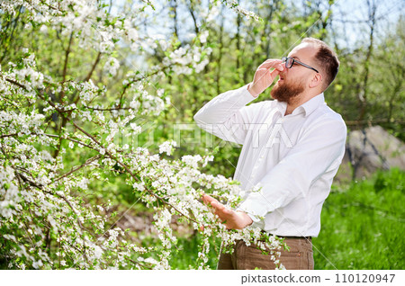 Man allergic suffering from seasonal allergy at spring in blossoming garden at springtime. Young man with glasses sneezing, closing nose by hand in front of blooming tree. Spring allergy concept 110120947