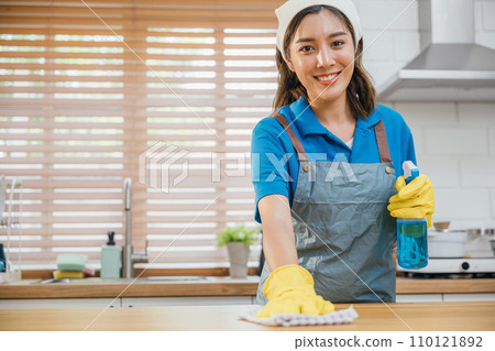 Asian woman in yellow gloves cleans wooden kitchen counter with liquid spray ensuring hygiene. Housekeeping service focusing on home cleanliness. Clean disinfect home care. maid household job. 110121892
