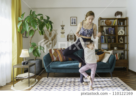 Young brown-haired instructor teaching yoga to a 4 year old girl in the living room 110122169