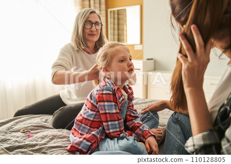 Little blonde girl sitting at the bed while her loving grandmother making hairstyle for her Little blonde girl sitting at the bed while her loving grandmother making hairstyle for her 110128805