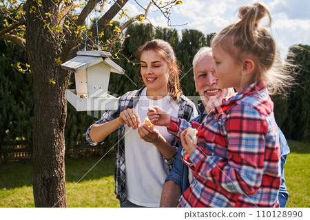 Joyful grandfather holding little girl and standing with his daughter near wooden birdhouse 110128990