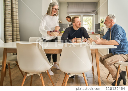 Cheerful senior woman serving table while her son talking with father Cheerful senior woman serving table while her son talking with father 110129464