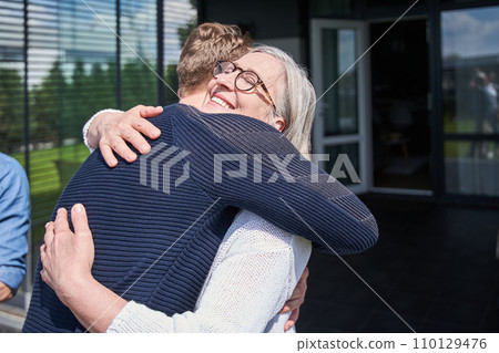Smiling senior woman embracing her adult son while standing near the house Smiling senior woman embracing her adult son while standing near the house 110129476