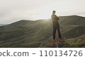 An adult young man in a suit and sneakers stands on a stone with his arms crossed on his chest and admires the sunset in the mountains. Unique landscape. Back view. Slow motion 110134726