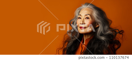 Smiling, elderly, gorgeous Asian woman with gray long hair and perfect skin, on an orange background, banner. 110137674