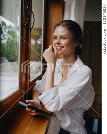Beautiful woman with phone in hand standing at home window, freelancer on vacation, home convenient lifestyle with online work, cyber security concept, smart home. Beautiful woman with phone in hand standing at home window, freelancer on vacation, home convenient lifestyle with online work, cyber security concept, smart home. 110140836