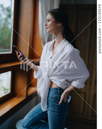 Young woman standing at the window with phone in hand smiling and looking out the window at nature, spring mood, home simple clothes, home cozy atmosphere, aesthetic lifestyle. Young woman standing at the window with phone in hand smiling and looking out the window at nature, spring mood, home simple clothes, home cozy atmosphere, aesthetic lifestyle. 110140839