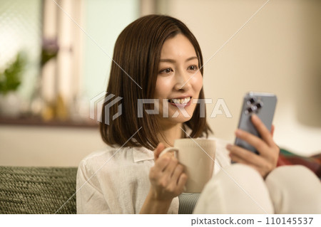 Middle-aged woman relaxing on the sofa, drinking coffee and looking at her smartphone 110145537