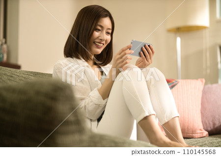 Middle-aged woman relaxing on the sofa and looking at her smartphone Middle-aged woman relaxing on the sofa and looking at her smartphone 110145568