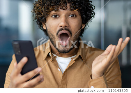 Close-up portrait of a young Indian man looking shocked and surprised at the camera while holding a mobile phone and spreading his hands. Close-up portrait of a young Indian man looking shocked and surprised at the camera while holding a mobile phone and spreading his hands. 110145805