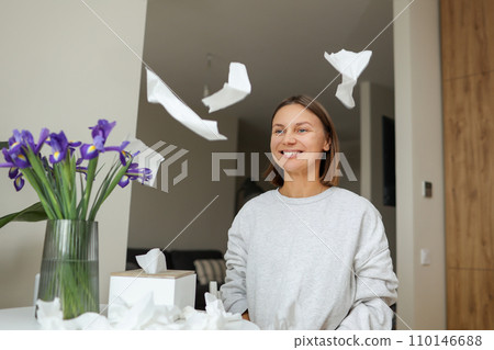 Seasonal allergy free. Happy young woman throws tissues up at home. Healthy life without of runny nose, itching or cough symptoms. Allergen bouquet of iris flowers at vase and used napkins on table. 110146688