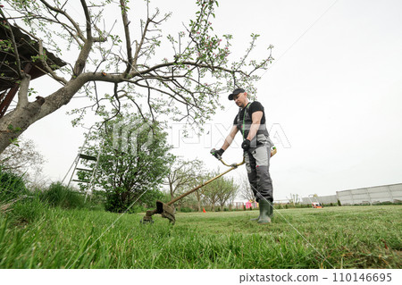 Side view of professional gardener in protective apparel is trimming green grass. Man worker mows weeds with hand electric or petrol lawn trimmer, mower in city park or backyard. Gardening care tools Side view of professional gardener in protective apparel is trimming green grass. Man worker mows weeds with hand electric or petrol lawn trimmer, mower in city park or backyard. Gardening care tools 110146695