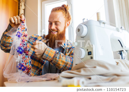 handsome redhaired man with long beard sews at a sewing machine at home studio 110154723