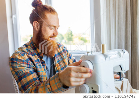 handsome redhaired man with long beard sews at a sewing machine at home studio handsome redhaired man with long beard sews at a sewing machine at home studio 110154742