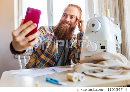 handsome redhaired man with long beard sews at a sewing machine at home studio handsome redhaired man with long beard sews at a sewing machine at home studio 110154744