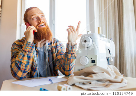 handsome redhaired man with long beard sews at a sewing machine at home studio handsome redhaired man with long beard sews at a sewing machine at home studio 110154749