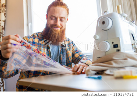 handsome redhaired man with long beard sews at a sewing machine at home studio handsome redhaired man with long beard sews at a sewing machine at home studio 110154751